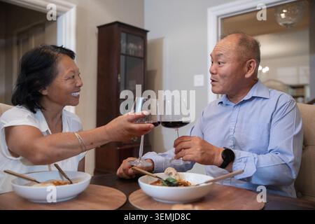 Una coppia cinese senior che porta bicchieri da vino rosso al tavolo da pranzo di casa con ciotole bianche e bacchette Foto Stock