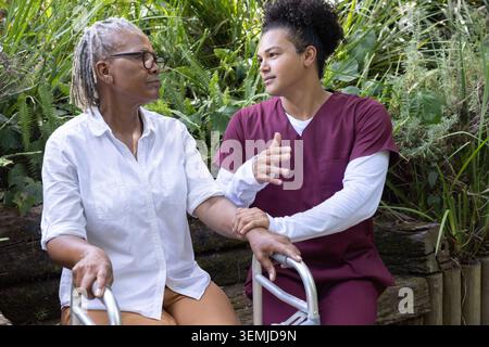 Caregiver inginocchiato in scrub di color marrone tenendo in mano una donna afroamericana anziana con un braccio in giardino Foto Stock