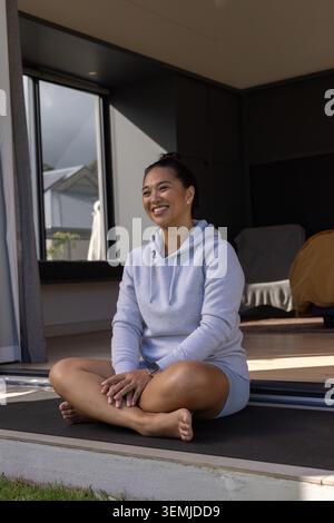 Donna asiatica di mezza età seduta a gambe incrociate sul tappetino all'ingresso, con felpa con cappuccio grigia e smartwatch Foto Stock