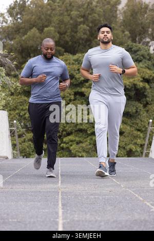 Due amici maschi che fanno jogging fianco a fianco su un sentiero pavimentato del parco, indossando scarpe da running e orologi Foto Stock