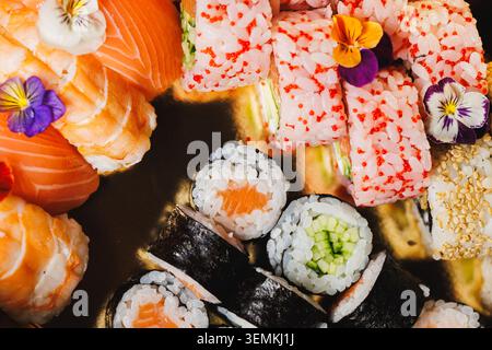 Una selezione di sushi roll, sashimi e colorati fiori commestibili è disposta su un piatto nero. Il sushi include ripieni e condimenti diversi. Il Foto Stock