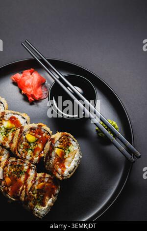 Un piatto mostra i sushi roll disposti ordinatamente su un piatto nero. La salsa di soia si trova in una piccola ciotola accanto allo zenzero sottaceto e al wasabi. Le bacchette poggiano su t Foto Stock