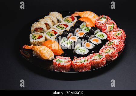 Un piatto nero rotondo può contenere diversi tipi di sushi. Ci sono panini con pesce, verdure e riso. Il piatto presenta un mix di colori e forme, Foto Stock