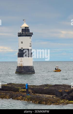 14 maggio 2026, angleseyTrwyn Du Lighthouse a Penmon Point, Anglesey, con le sue strisce bianche e nere con una piccola barca gialla sul mare e un maschio Foto Stock