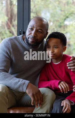 Padre-figlio maschio afroamericano seduto a casa su un sedile in pelle vicino a una grande finestra, tenendo la spalla Foto Stock