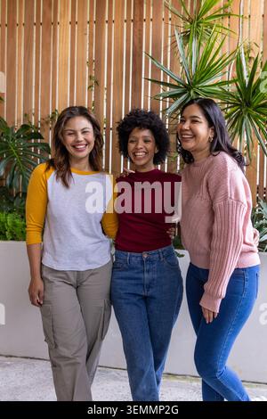 Diverse amiche si alzano e sorridono sul patio in cemento da una piantatrice rialzata e da un muro di legno Foto Stock