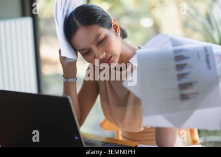 Donna d'affari stressata che esamina documenti finanziari e laptop in un bar dell'ufficio, mostra pressione contabile, problemi di budget, sovraccarico di documenti cartacei, a. Foto Stock