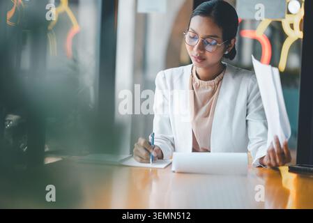 Donna d'affari concentrata che scrive appunti e tiene i documenti alla scrivania, esamina i documenti, pianifica le attività e organizza le informazioni durante un periodo di calma Foto Stock
