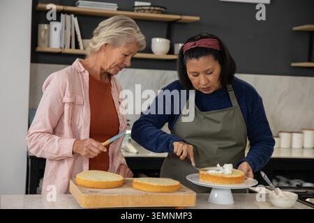 Donne anziane che diffondono glassa bianca sul banco per torte e strati di spugna al bancone di casa, in grembiuli Foto Stock