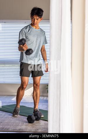 Uomo asiatico che esegue bicep curl tenendo il manichino esagonale sul tappetino verde per esercizi a casa in abbigliamento sportivo Foto Stock