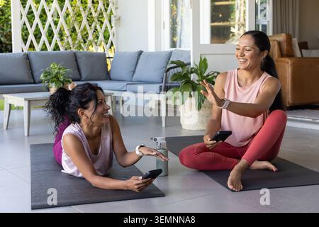 Diverse amiche in abbigliamento sportivo si rilassano sui tappetini da yoga nel patio con il treppiede per biberon del telefono Foto Stock