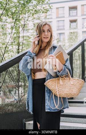 Una giovane donna parla sullo smartphone mentre cammina giù per le scale della città tenendo un laptop. Lavorare in movimento, creatività indipendente, lavoro remoto flessibile, moderno Foto Stock