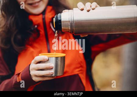 La donna sorridente versa la bevanda calda dai termos metallici nella tazza all'aperto. attività ricreative stagionali nella foresta, abbigliamento da campeggio funzionale, trekking con clima freddo Foto Stock