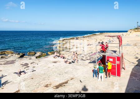 Sliema, Malta - 16 maggio 2025: Bagnini sorvegliano i nuotatori e i bagnanti da una torre rossa lungo la costa rocciosa di Sliema Beach con un sole luminoso Foto Stock