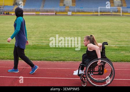 Atleta in sedia a rotelle che si allena sulla pista da corsa con Coach che cammina accanto a lei nello stadio Foto Stock