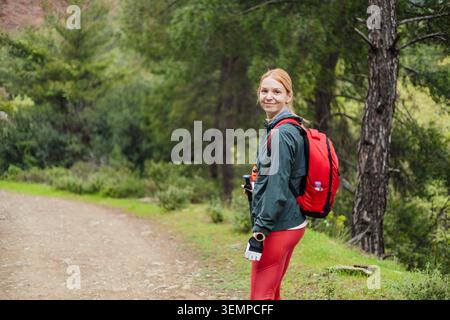 Donna sorridente con uno zaino rosso che fa trekking in una foresta Foto Stock
