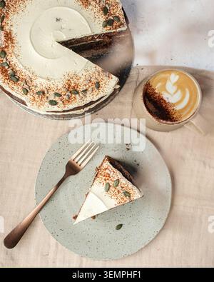 Un bicchierino piatto di una torta di carote affettata con glassa di formaggio cremoso, una fetta di torta su un piatto con una forchetta e un cappuccino caldo con la Foto Stock