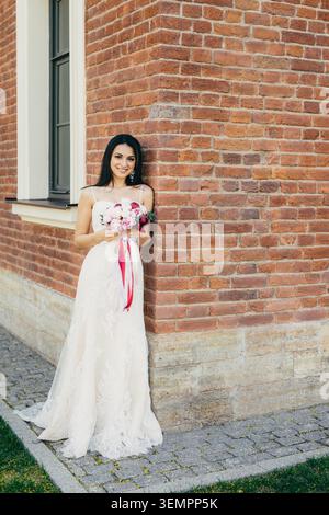 Abito da sposa in pizzo bianco appoggiato su una parete di mattoni rossi che regge un bouquet di fiori rosa e Bordeaux Foto Stock