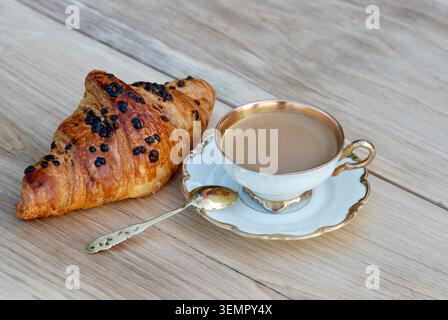 Caffè in antico set da caffè con cucchiaio placcato oro argento su piattino e croissant su tavolo in quercia. Vista frontale ravvicinata. Foto Stock