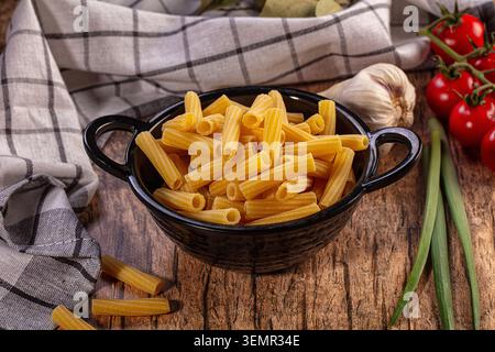 Maccheroni crudi in ciotola nera, fondo rustico in legno. Ingredienti freschi, pomodori di vite, aglio, cipolle verdi, foglie di alloro, asciugamano a scacchi. Natur Foto Stock