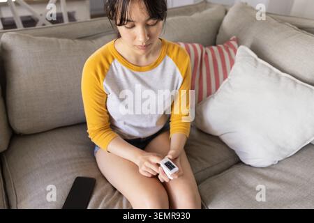 Donna asiatica seduta su un divano grigio chiaro a casa mentre controlla il pulsossimetro a punta di dita vicino allo smartphone Foto Stock
