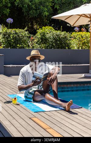 Uomo afro-americano seduto sul ponte della piscina, asciugamano da lettura, libro cartaceo con cappello di paglia e occhiali da sole Foto Stock