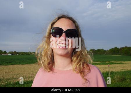 Donna con lunghi capelli biondi e occhiali da sole, sorridente in un ambiente esterno. Sta indossando un top rosa e sembra felice. Lo sfondo mostra una l. Rurale Foto Stock