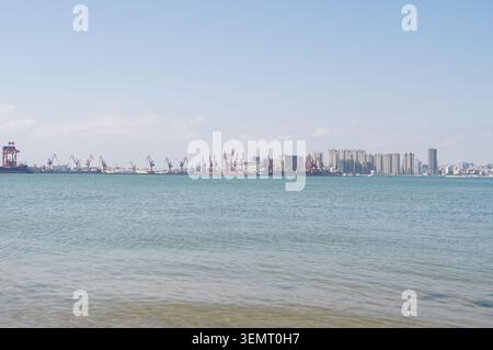 Paesaggio costiero di Zhanjiang, Mar Cinese meridionale, Guangdong, vecchie barche da pesca in legno, palafitte sulla spiaggia e distanti terminal portuali per container Foto Stock