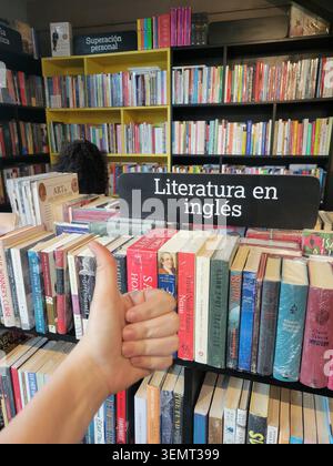 Una vista ravvicinata della mano di una persona che si avvicina ai libri sugli scaffali alti all'interno di una libreria affollata, con file di titoli vari e un libro cartaceo colorato Foto Stock