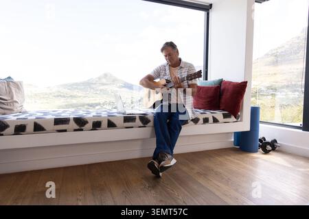 Uomo maturo seduto sulla panchina di casa che suona la chitarra acustica con laptop, rullo in schiuma, manubri Foto Stock