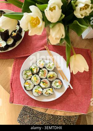 Gli involtini di sushi sono disposti ordinatamente su un piatto bianco. I panini contengono cetriolo, pesce rosso e uova. Sono serviti con bacchette su un tappeto blu e verde Foto Stock