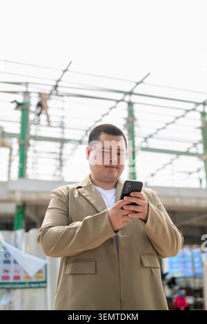 Una ripresa ad angolo basso di un uomo d'affari asiatico medio adulto della regione di Isan che usa il suo telefono sotto un progetto di costruzione in acciaio Foto Stock