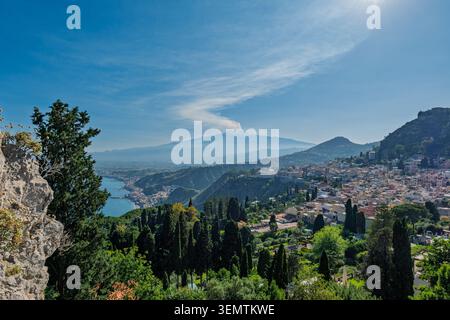 Vista panoramica sopraelevata di Taormina, storica cittadina collinare in Sicilia, Italia, con l'iconica silhouette dell'Etna che emette un pennacchio di fumo visibile Foto Stock