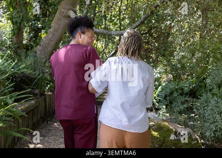 Assiste diverse persone in scrub di marrone che guidano una donna afroamericana anziana lungo il sentiero di ghiaia con felci Foto Stock