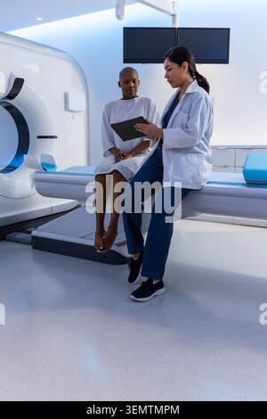 Paziente donna vestita seduto con fornitore asiatico femminile in scrub revisione tablet a CT Foto Stock