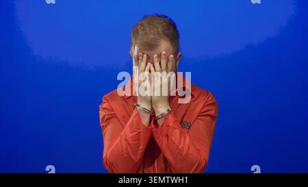 Tuta da giovane in prigione arancione con mani che coprono il viso e manette in metallo in studio contro il muro blu; rimorso. Foto Stock