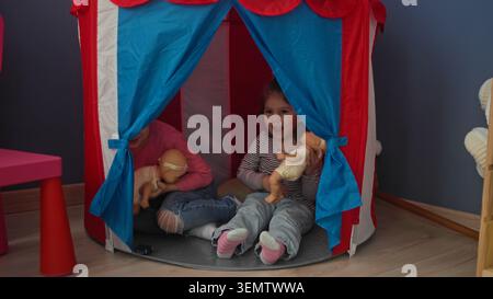 Due bambine tengono le bambole e si siedono all'interno di una tenda da da gioco rossa e blu in una stanza da gioco domestica, una sorridente e una in bocca; gioia d'infanzia. Foto Stock