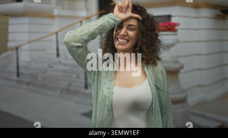 Donna che fa il segno con mano a fronte sui gradini di costruzione all'aperto, sorriso ampio che mostra i denti; giocosa. Foto Stock
