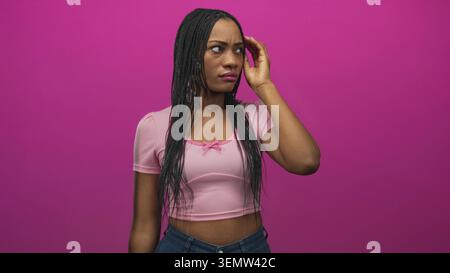 Donna che tocca i capelli intrecciati con la mano, jeans esposti e jeans in denim visibili in studio con sfondo rosa e collana visibile; contemplazione tranquilla. Foto Stock