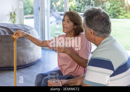 Coppia anziana seduta a casa, donna anziana che tiene in mano canne di legno e un bicchiere d'acqua Foto Stock