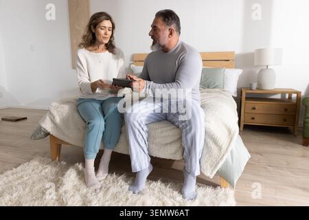 Coppia donna di mezza età e uomo maturo seduto sul letto condividendo un tablet in una camera da letto con luce soffusa Foto Stock