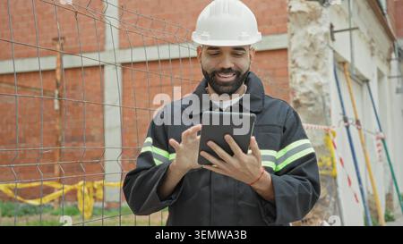 Man Architect in elmetto e giubbotto riflettente punta le dita sul tablet e tocca lo schermo della recinzione del cantiere, pianificando la sicurezza. Foto Stock