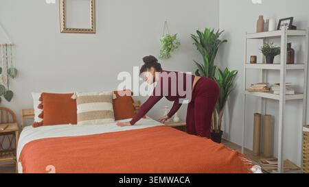 Donna che nasconde una coperta arancione sul letto con le mani e i cuscini liscianti mentre si piega vicino allo scaffale in una camera da letto dell'edificio; calma routine. Foto Stock