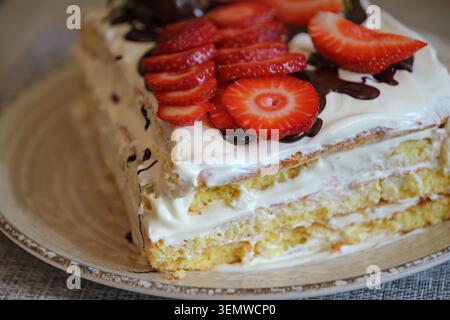 Vista ravvicinata di una pan di Spagna fatta in casa con glassa cremosa, fette di fragola fresche e gocciolatine di cioccolato. Dessert appetitoso servito su una ce Foto Stock
