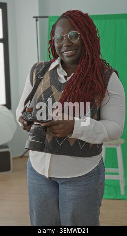 La donna tiene in mano la fotocamera e sorride mentre regola l'obiettivo in uno studio fotografico luminoso, indossa occhiali rotondi e un gilet argyle; creativo j Foto Stock