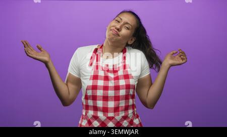 Una donna cuoca stringendo le palme e sorridendo nello studio viola indossando grembiule a scacchi rossi e tee bianco; giocosa incertezza. Foto Stock