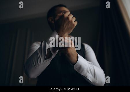 L'uomo regola il bracciale con un'illuminazione soffusa e intima. Concentrato, calmo e deliberato, la sua mozione suggerisce la preparazione. Ombre morbide incorniciano mani e camicia Foto Stock