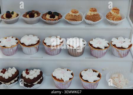 File di delicati cupcake con glassa e frutti di bosco. I vassoi bianchi ospitano prelibatezze con texture morbide e guarnizioni colorate. Ogni dessert è uniquel Foto Stock