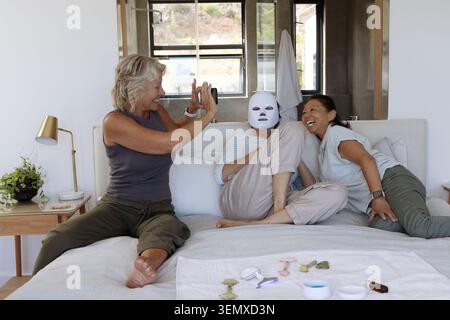 Donne diverse con anziane sedute sul letto che condividono la maschera e i rulli che scattano selfie con il telefono Foto Stock
