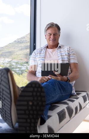 Uomo anziano seduto su una panchina disegnata da una grande finestra verticale, tenendo un tablet e mostrando suole Foto Stock